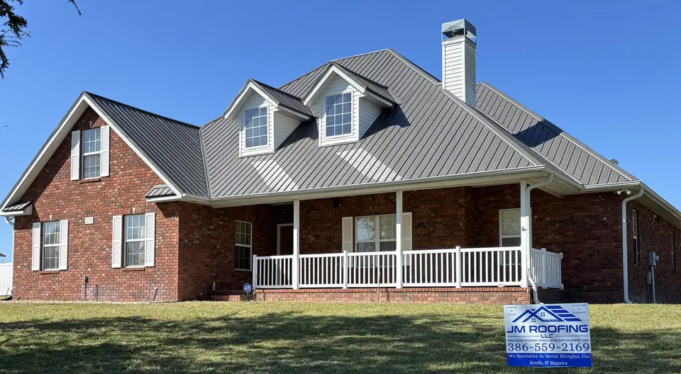 Brick house with dormers and a new gray standing seam metal roof, JM Roofing sign on the lawn